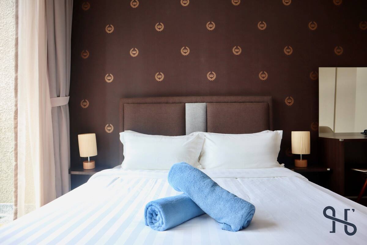 A hotel room with a large bed, two pillows, a rolled blue towel, a nightstand with a lamp, and a mirror on the wall.