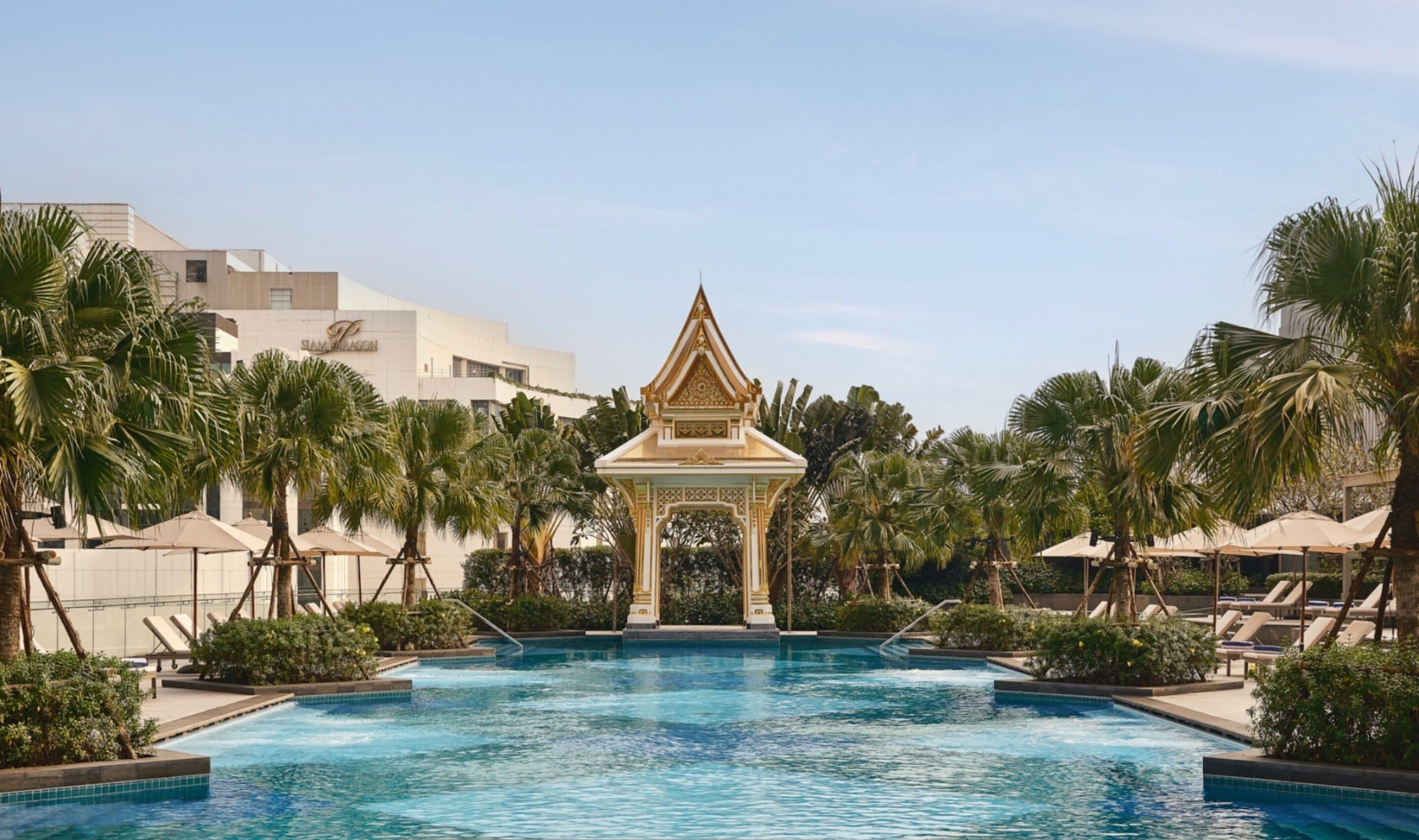 A swimming pool with a traditional pavilion in the center, surrounded by palm trees and lounge chairs.