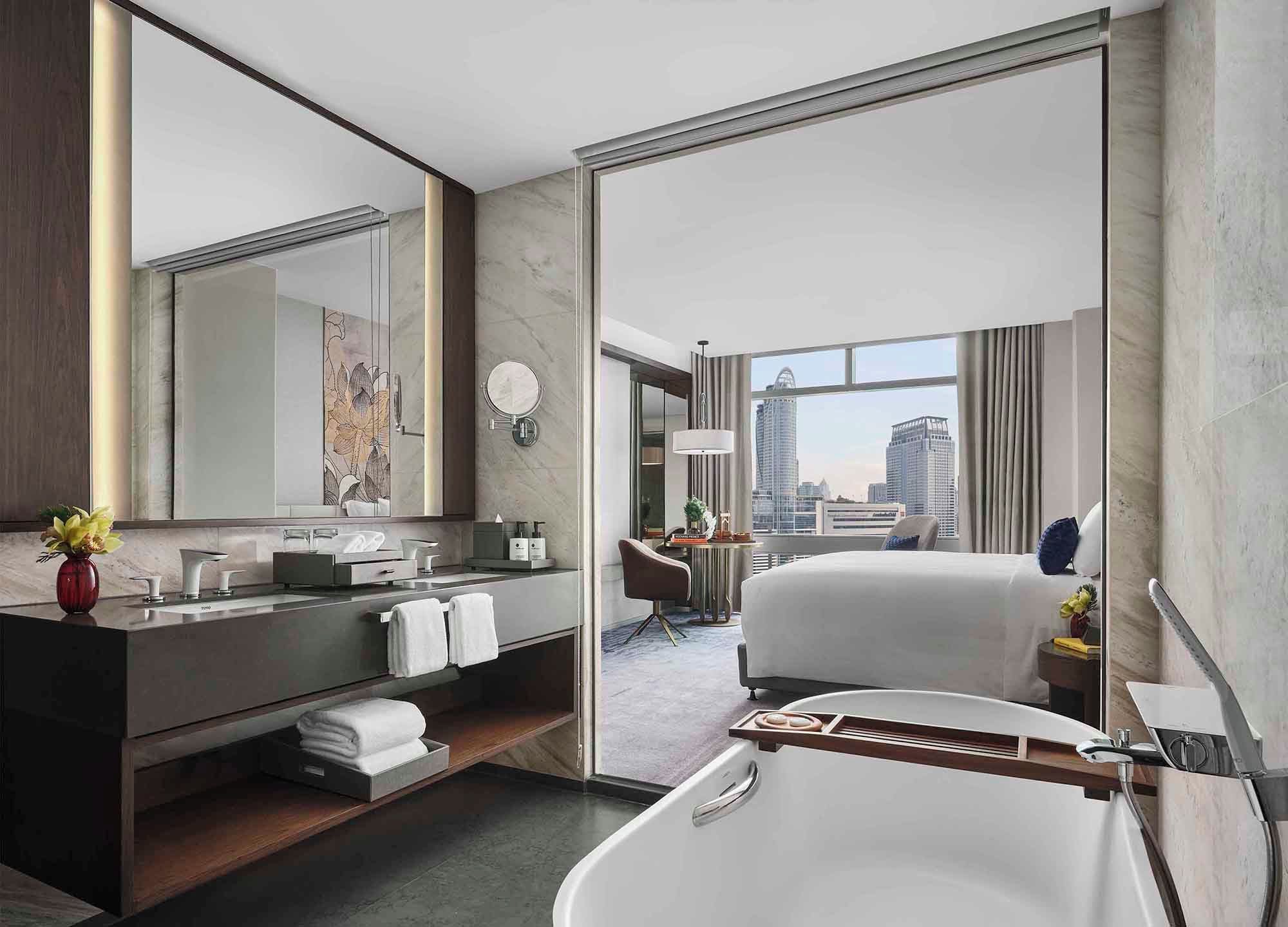 A modern hotel bathroom with a bathtub, a vanity with a mirror, and a city view through the window.
