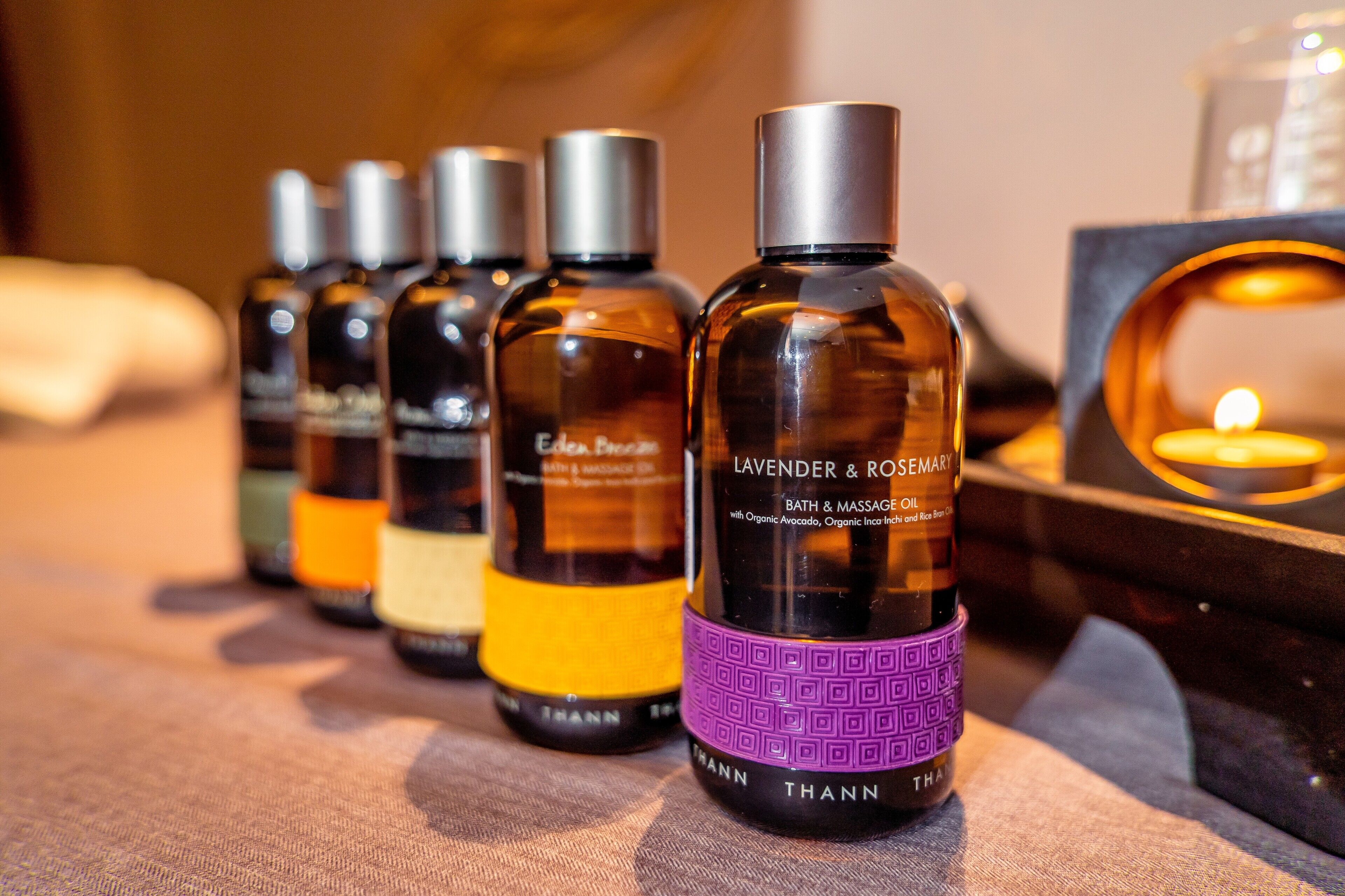 A row of bath and massage oil bottles with labels, arranged on a surface with a candle in the background.