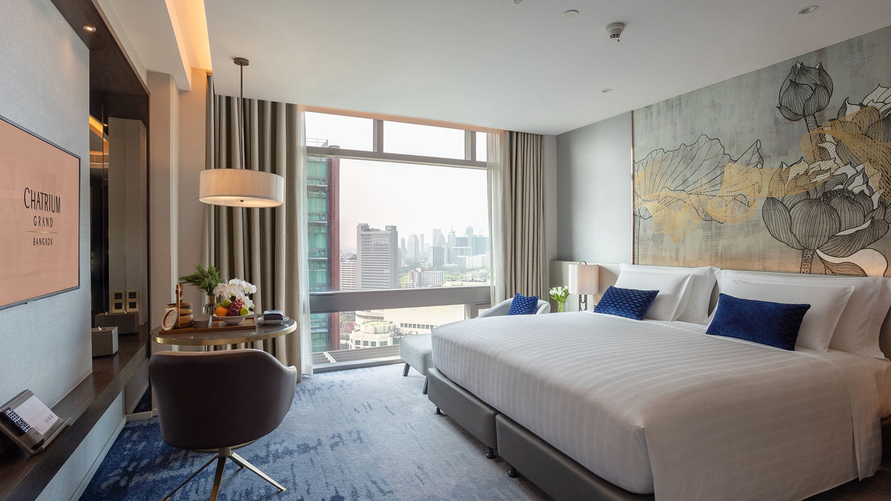 A hotel room with a large bed, a desk with a laptop, a chair, and a view of the city through large windows.