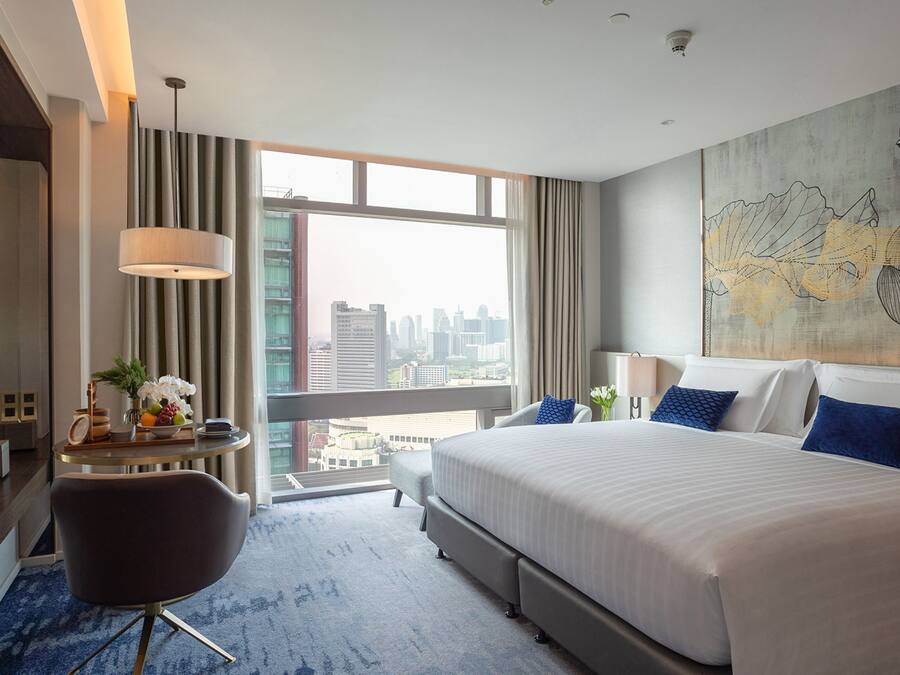 A hotel room with a large bed, a desk with a laptop, a chair, and a view of the city through large windows.