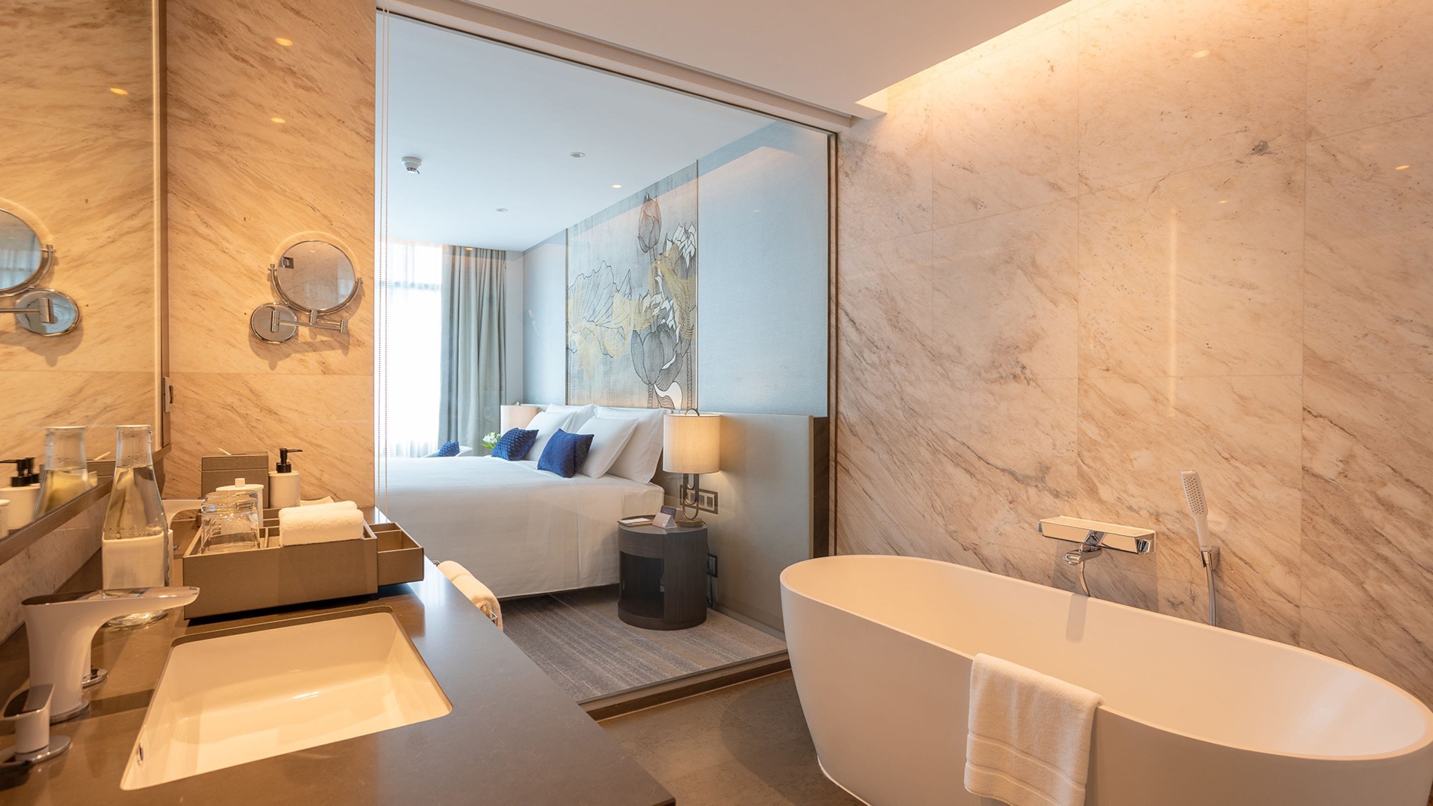 A modern hotel bathroom with a large bathtub, a separate shower area, a vanity with a mirror, and a bed with a view of the cityscape.