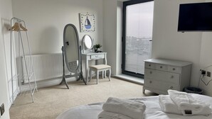 A bedroom with a bed, a vanity with a mirror, a radiator, a window with blinds, and a television on the wall.