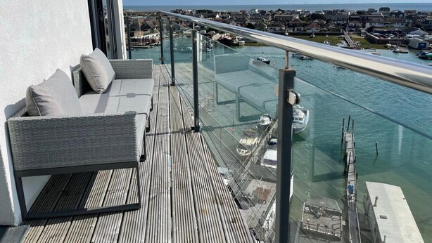 A balcony with a glass railing overlooking a marina with boats and a distant cityscape.