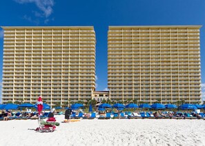 Ein Hotel mit mehreren Etagen und Balkonen, ein Strandbereich mit blauen Sonnenschirmen und ein Sandstrand.