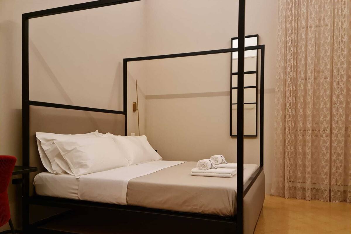 A four-poster bed with white linens and towels, a red chair, and a window with a floral curtain.
