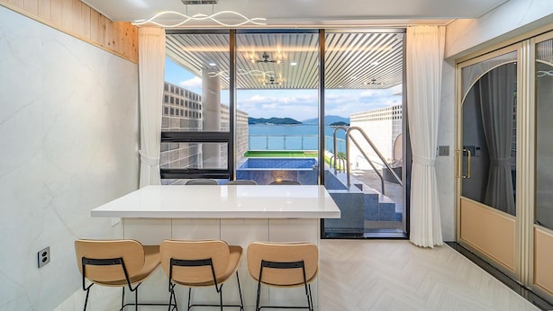 Ein modernes Zimmer mit einem weißen Tisch und drei Stühlen, eine Glastür zu einem Balkon mit Pool und Meerblick.