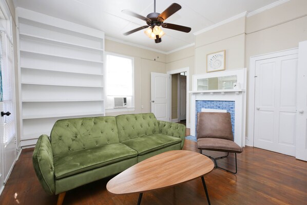 Una sala de estar con un sofá verde, una mesa de centro de madera, un sillón marrón, un ventilador de techo y una chimenea.