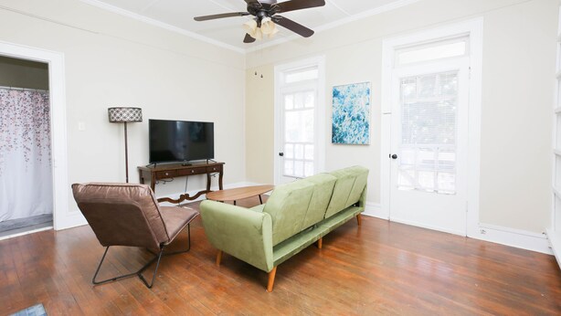 A living room with a green sofa, a brown chair, a wooden coffee table, a TV on a stand, and a ceiling fan.