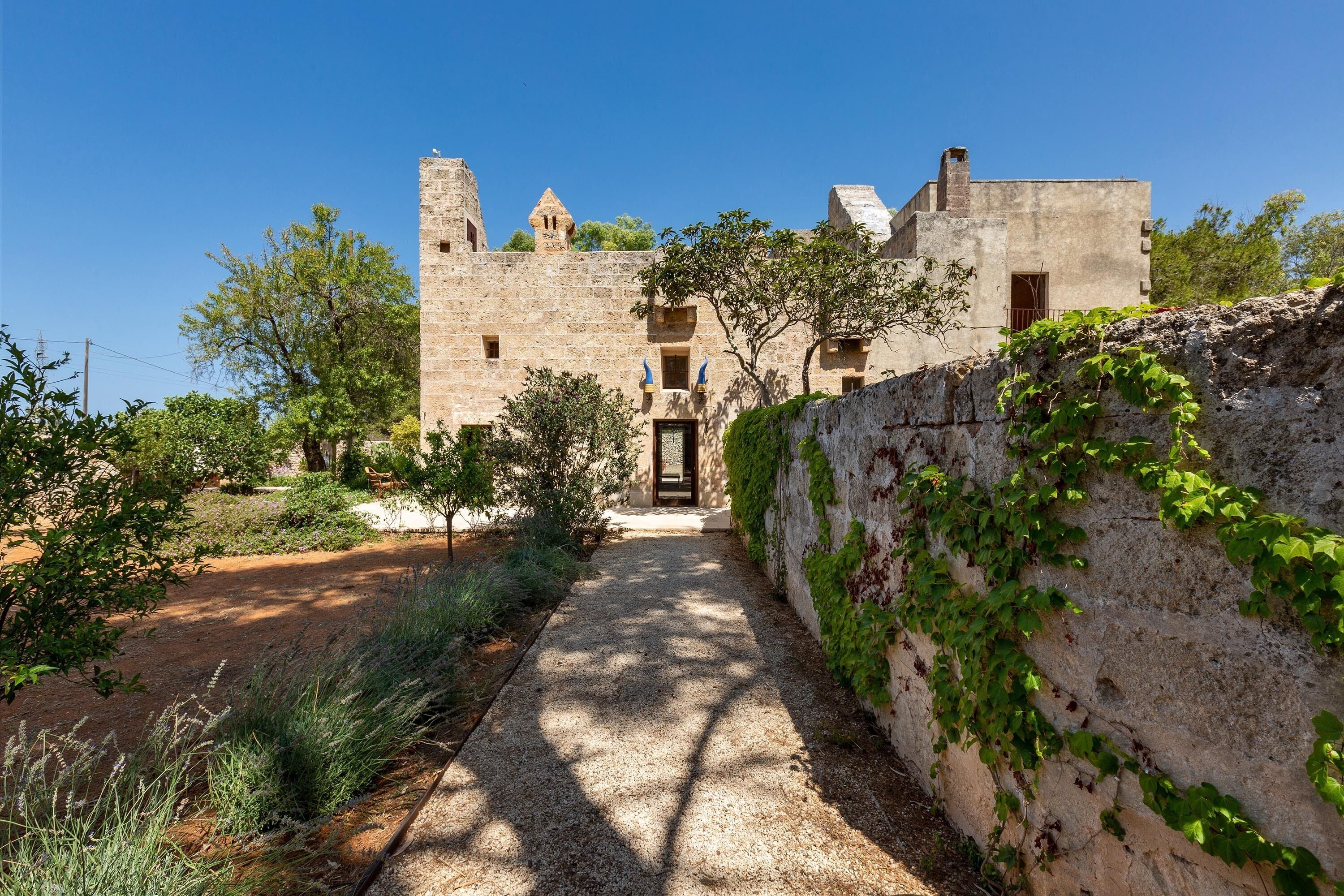 A stone building with a central door and windows, surrounded by greenery and a gravel path.