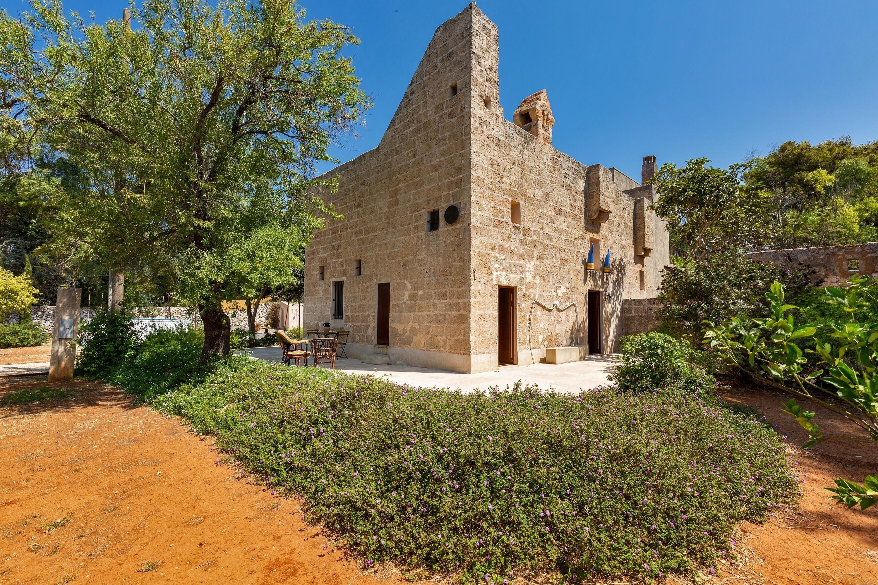 Un edificio de piedra con una torre destacada, rodeado de follaje y un sendero de tierra.
