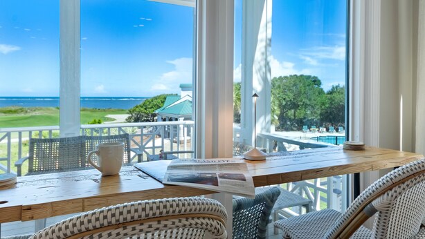 A wooden table with a newspaper, a white mug, and a wicker chair in a room with large windows overlooking a pool and a golf course.