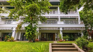 Un edificio de varios pisos con fachada blanca, balcones y un jardín bien cuidado con escalones que conducen a la entrada.