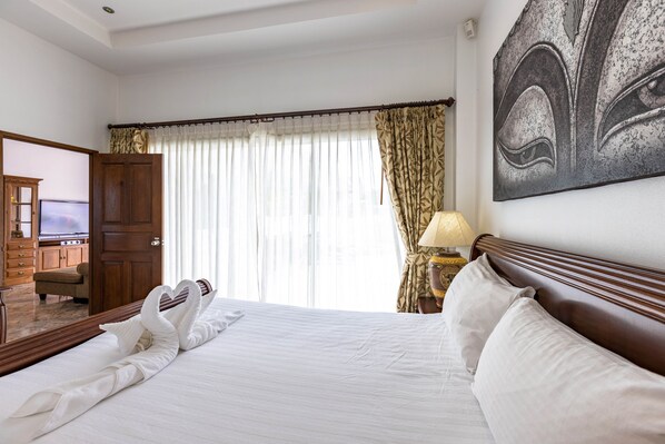 A hotel room with a large bed, a wooden headboard, a painting on the wall, a TV in the corner, and a window with curtains.