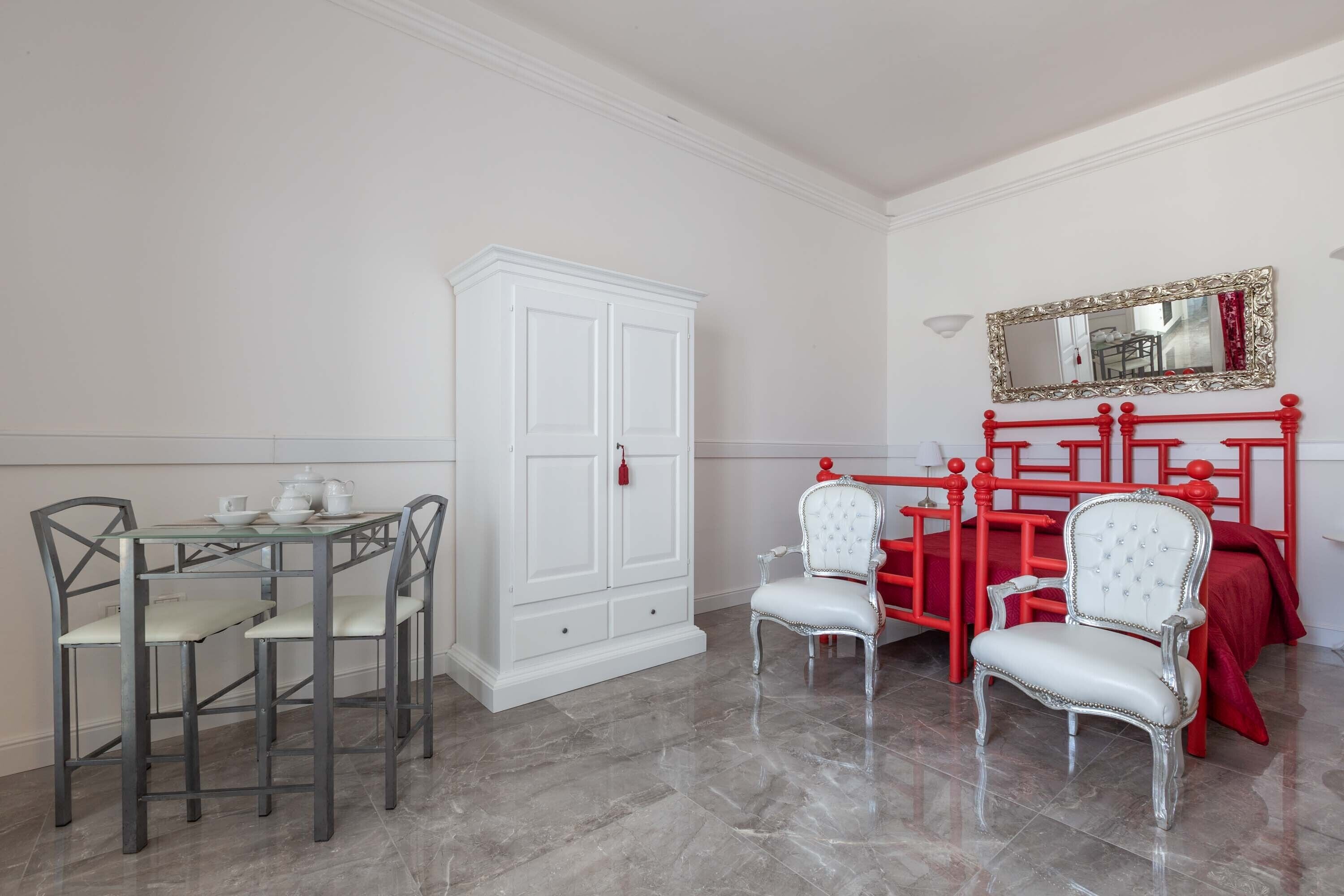A room with a dining table, chairs, a wardrobe, a bed with a red headboard, and a mirror on the wall.
