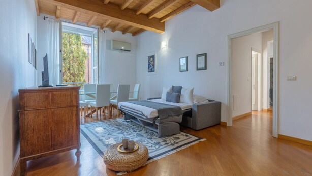 A bedroom with a bed, bedside tables, a wooden wardrobe, a dining area with chairs, and a view through a window.