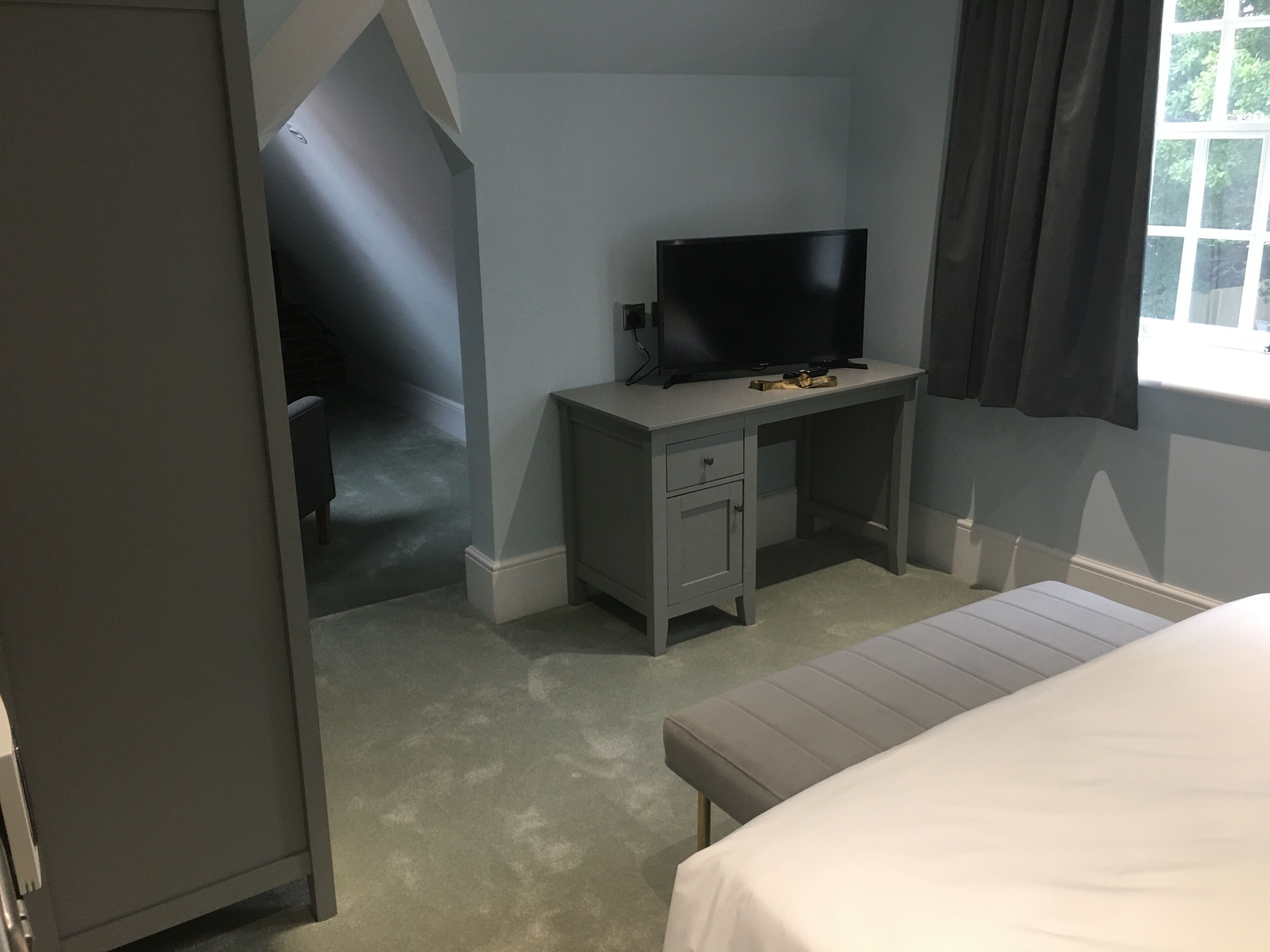 A bedroom with a bed, a desk with a TV, a window with curtains, and a door leading to another room.
