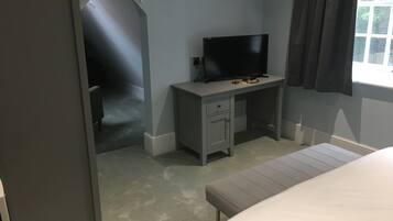 A bedroom with a bed, a desk with a TV, a window with curtains, and a door leading to another room.
