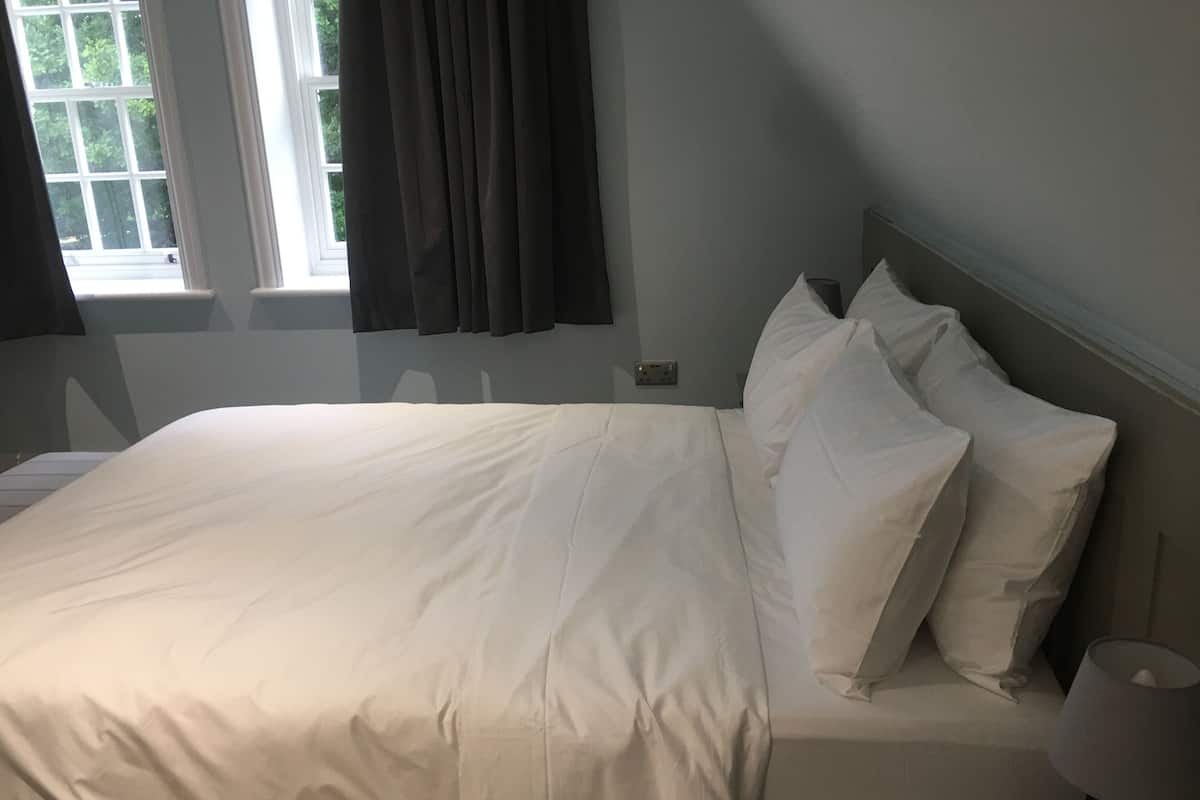 A neatly made bed with white linens, a bedside lamp, and a window with curtains.