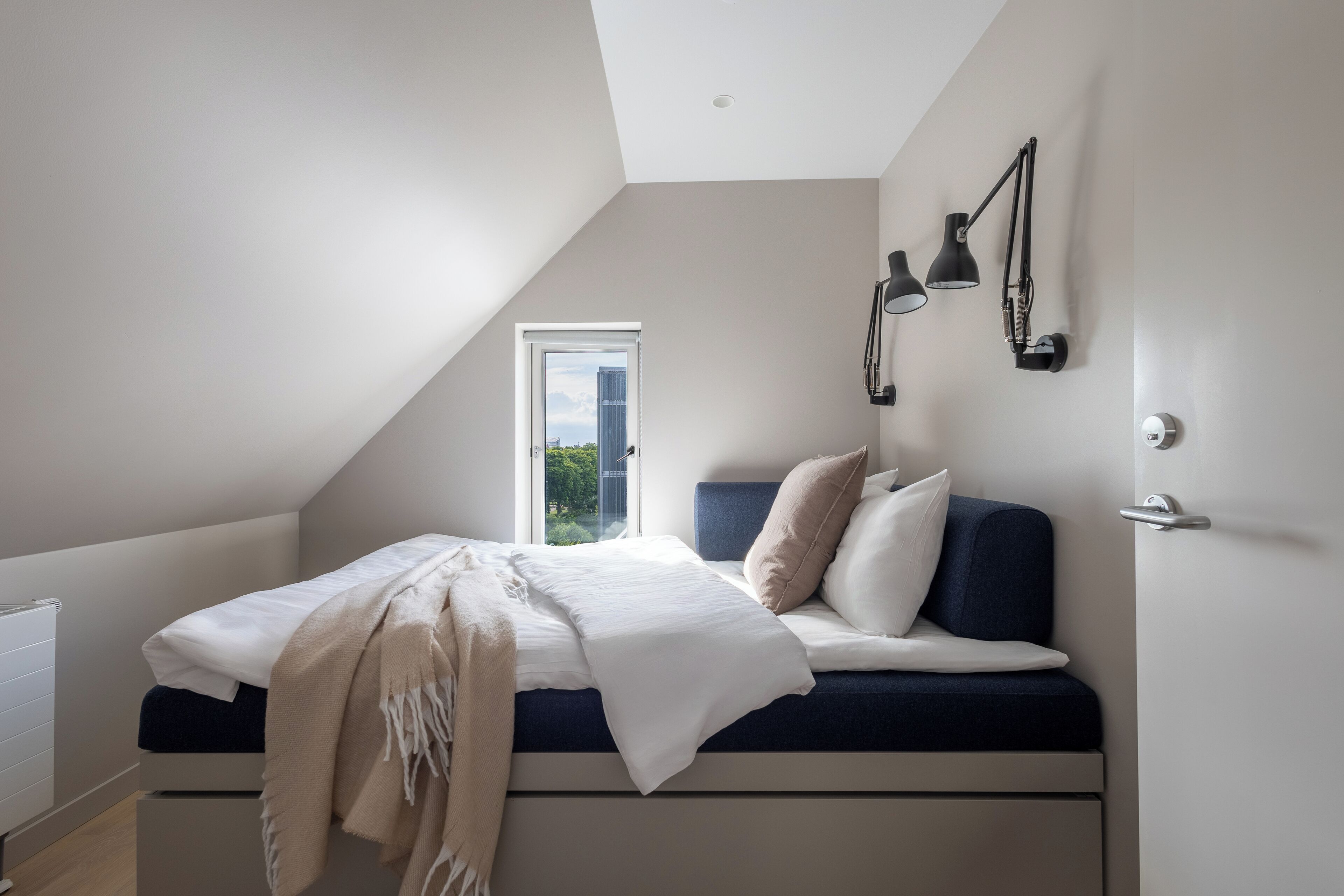Un dormitorio moderno con techo inclinado, una cama con ropa de cama blanca y azul, una manta color beige y dos lámparas de pared negras.