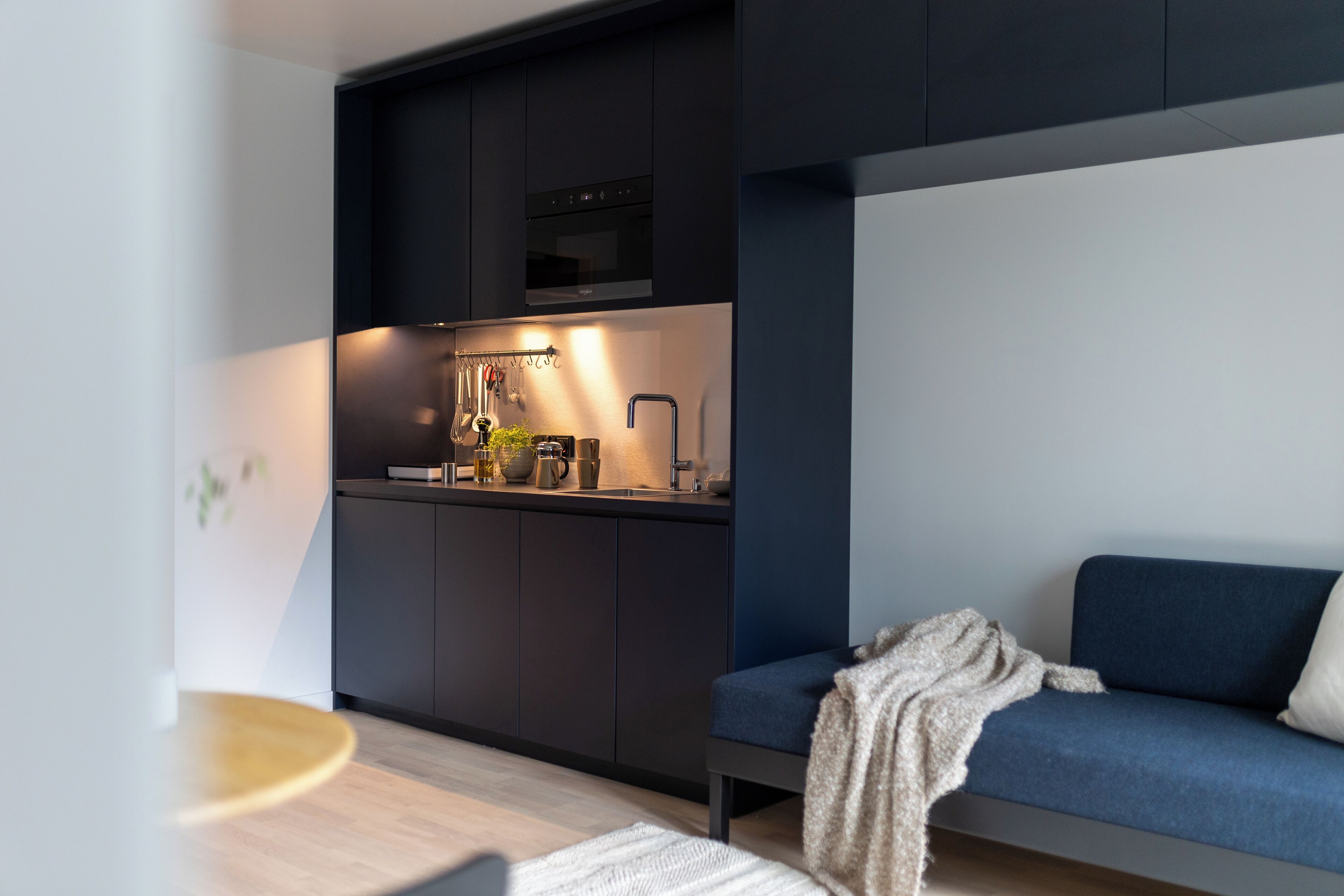 Eine moderne Küche mit dunklen Schränken, einem eingebauten Ofen und einem Essbereich mit einem blauen Sofa und einem kleinen runden Tisch.