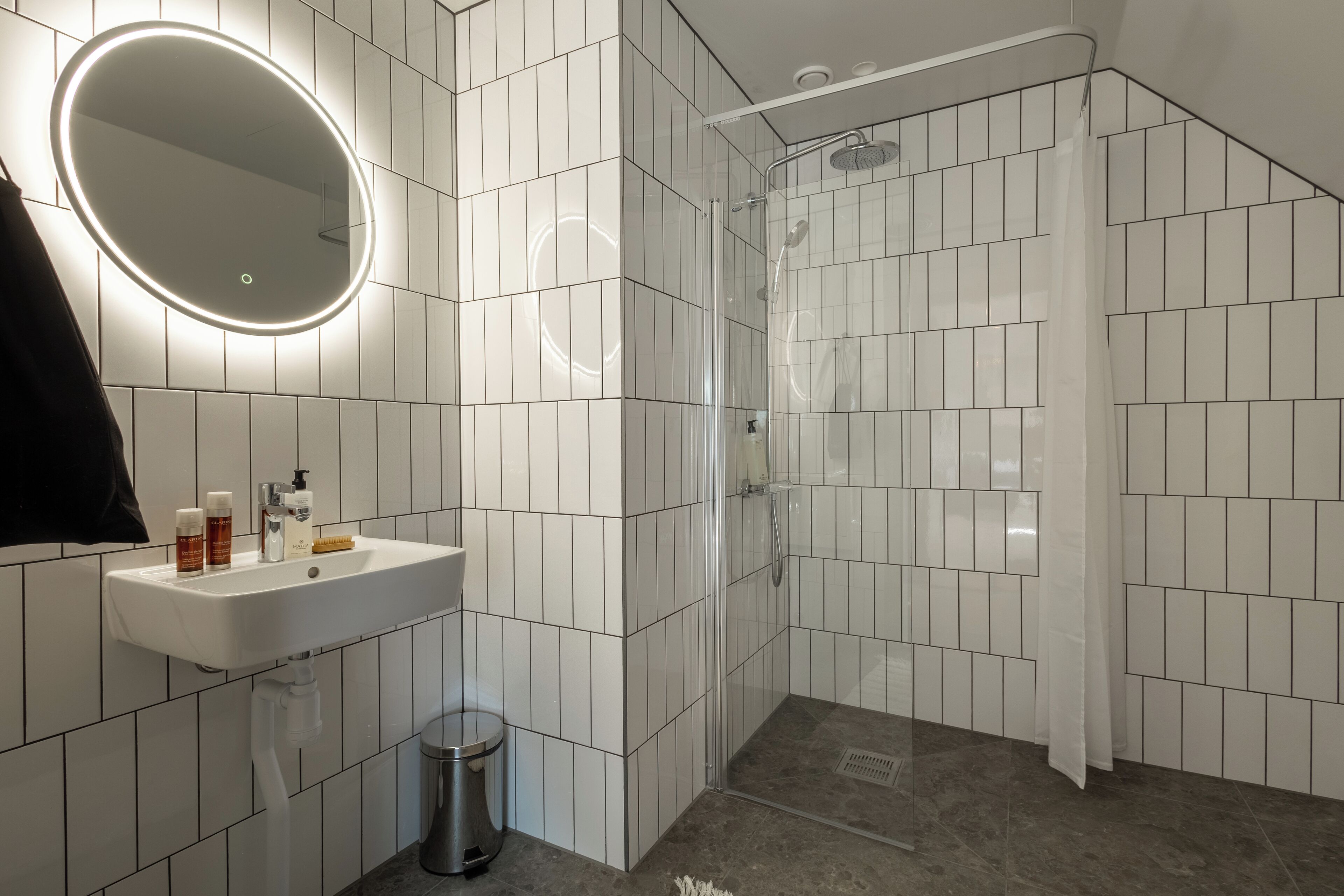 Een moderne badkamer met een witte betegelde muur, een ronde spiegel met verlichting, een wastafel met toiletartikelen en een douchehoek.