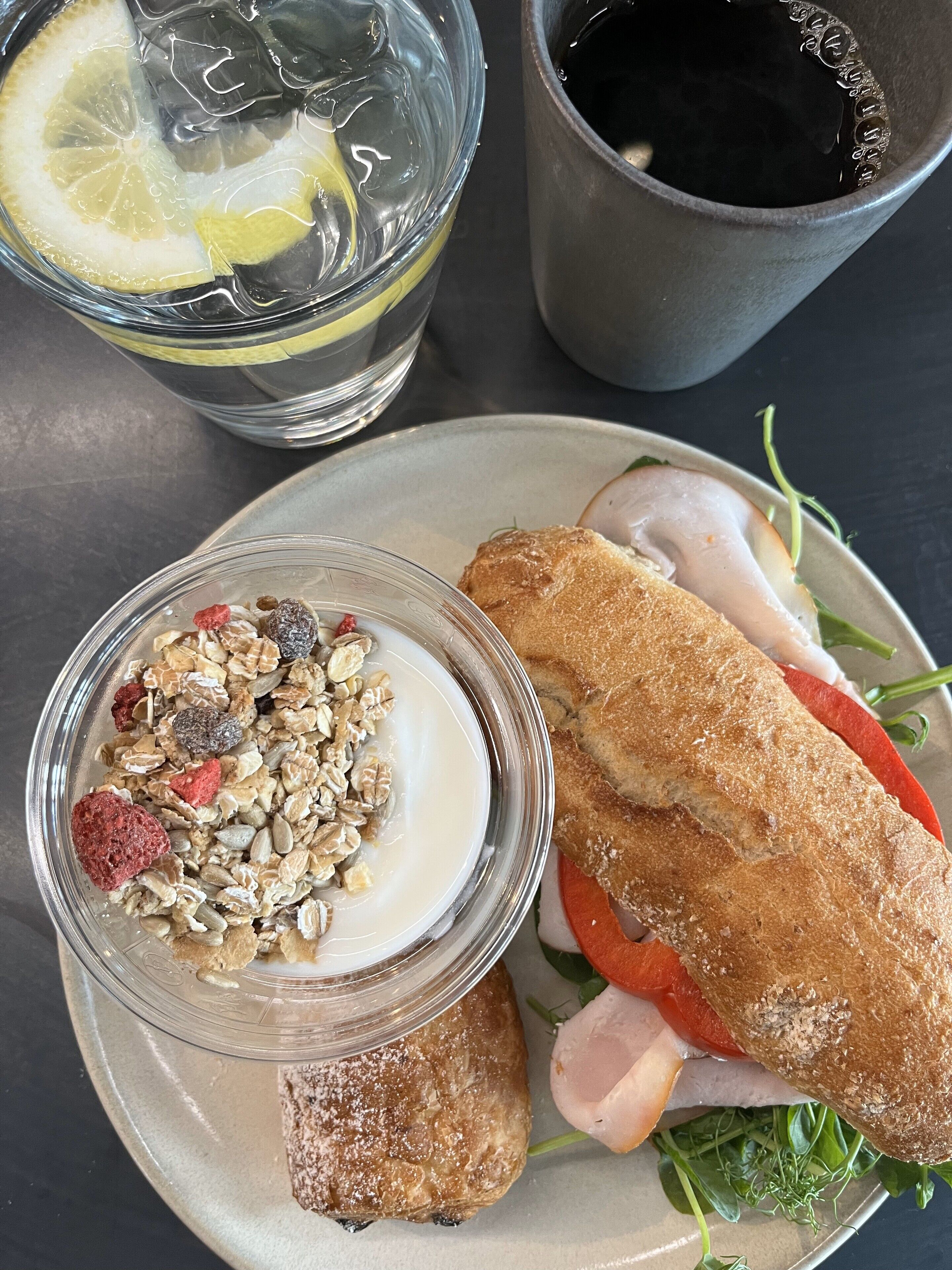 A meal consisting of a sandwich with tomato and leafy greens, a bowl of yogurt with granola and berries, and a glass of iced tea with a lemon slice.