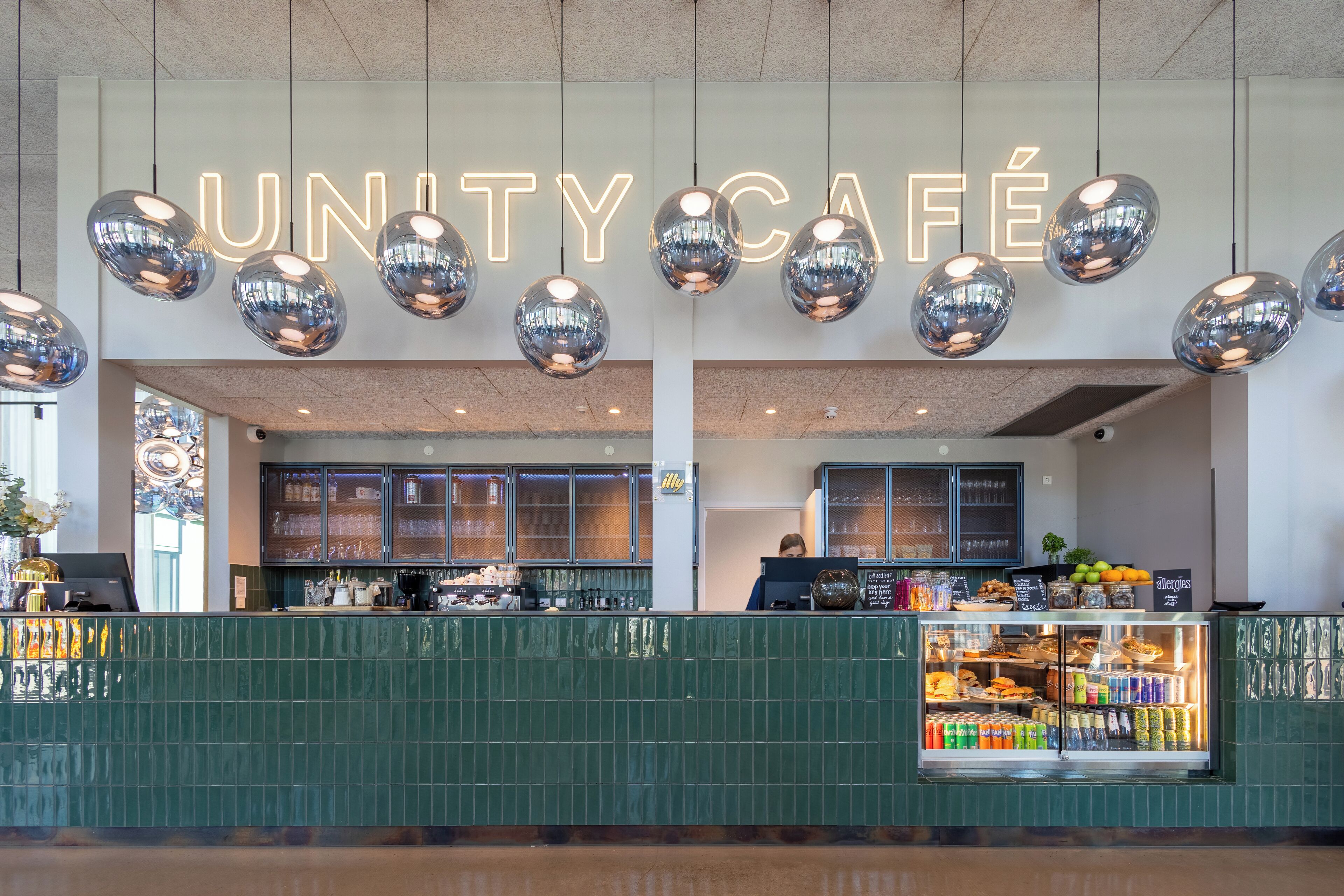 A café with a neon sign, hanging spherical lights, a counter with a display case, and a green tiled wall.
