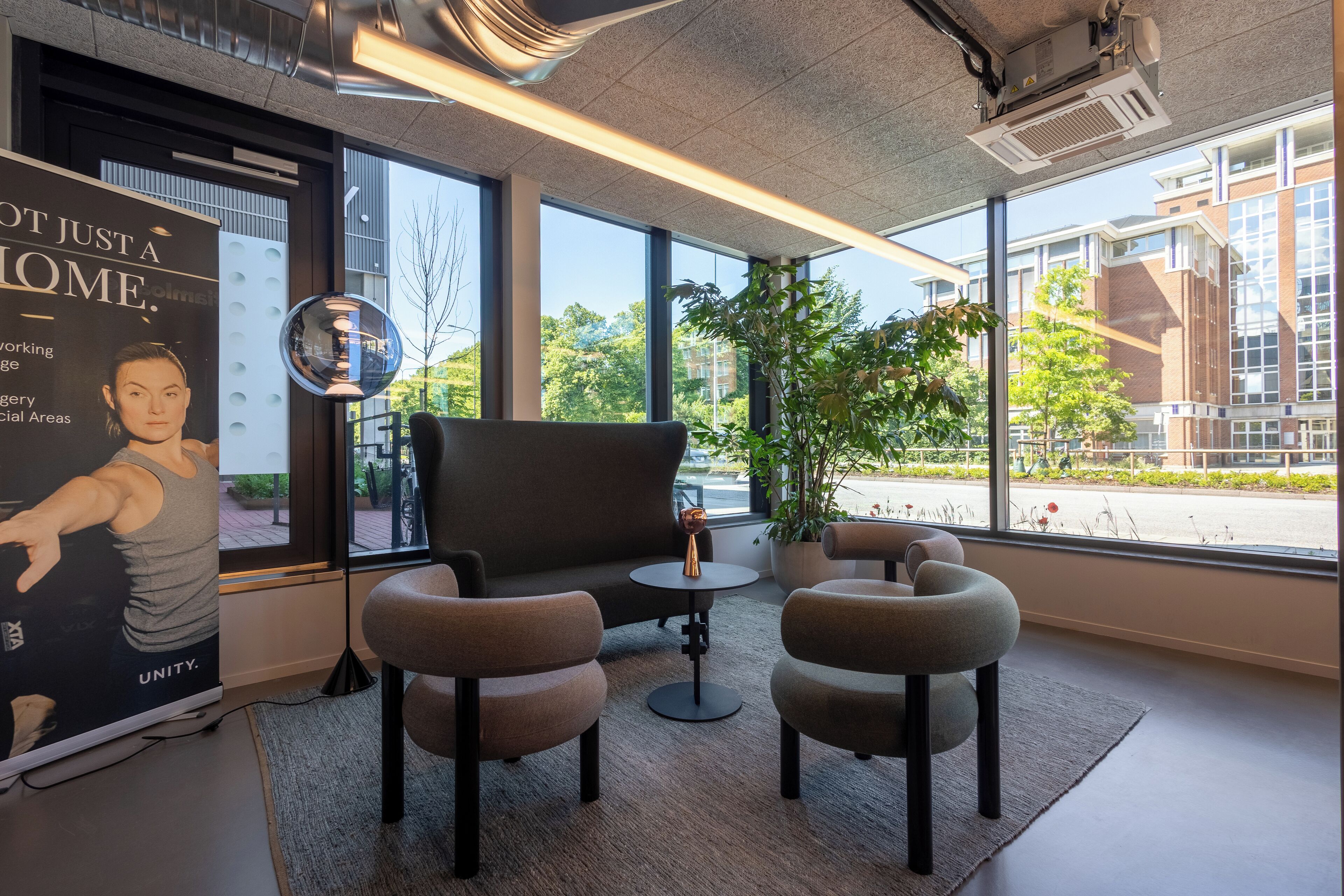 Un espacio de oficina moderno con ventanas amplias, un sofá negro, dos sillones, una mesita y una planta en maceta.