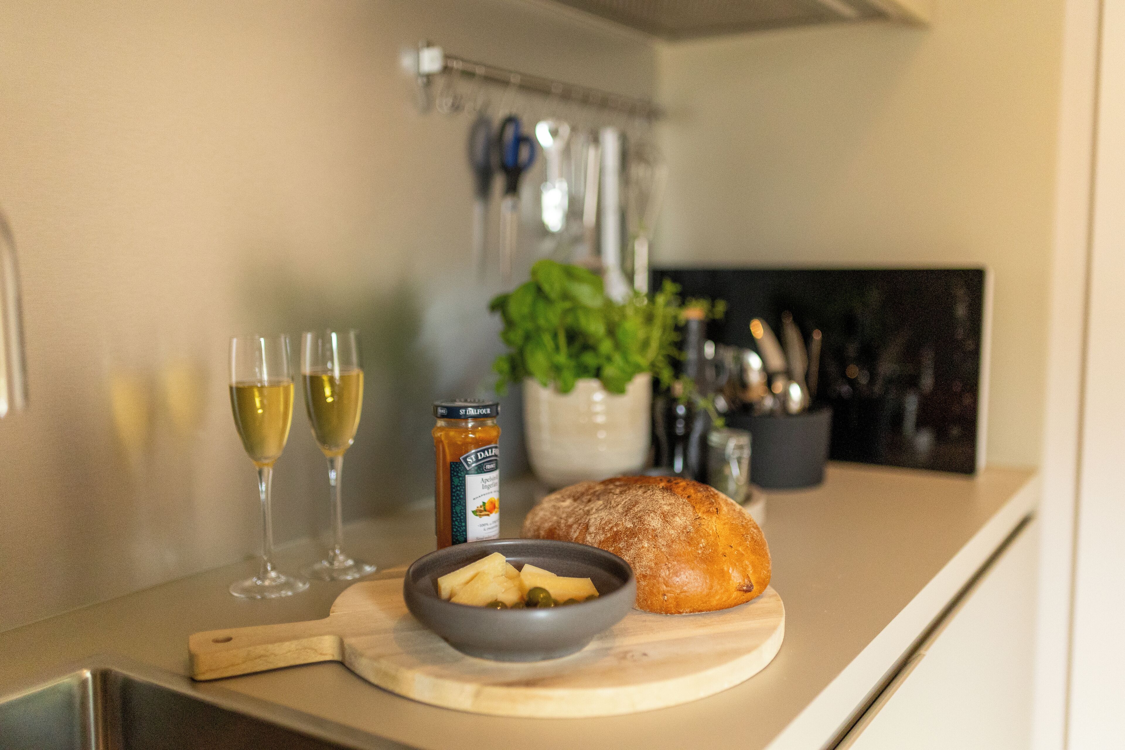 A kitchen counter with a loaf of bread, a bowl of fruit, and two glasses of wine.