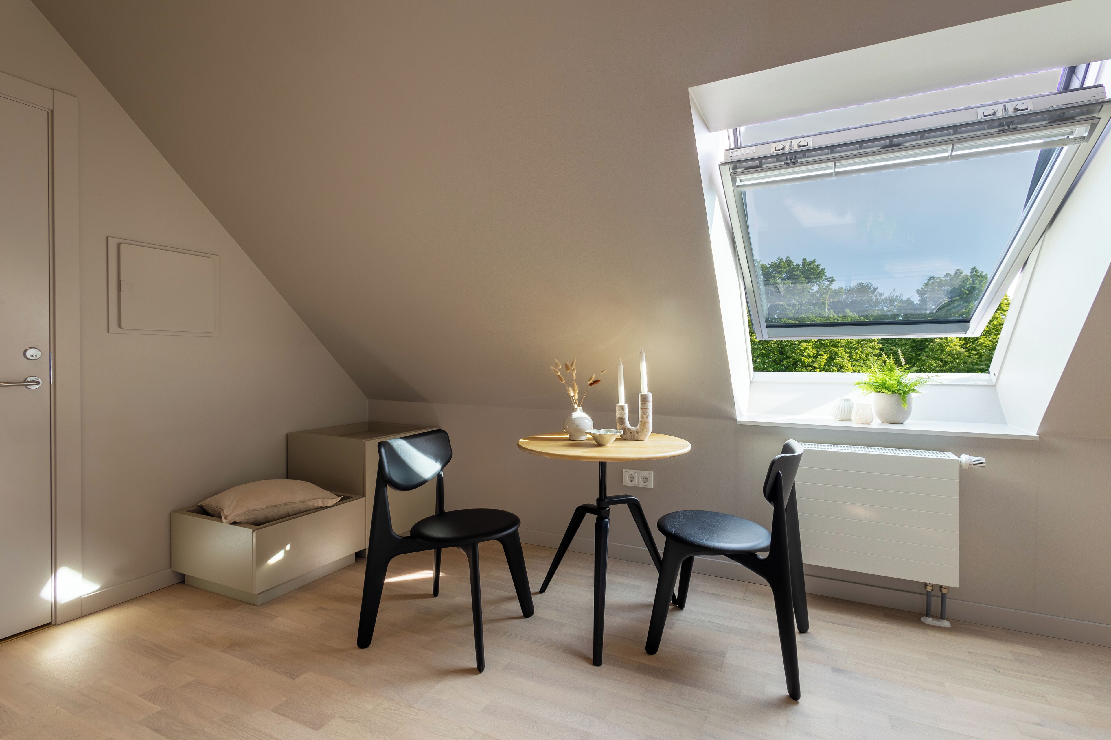 Ein kleiner, moderner Raum mit schrägem Dach, einer Dachfensterkuppel, einem runden Tisch mit Stühlen, einem Heizkörper und einem Bett mit Stauraum darunter.