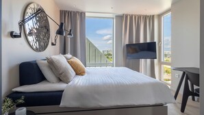 A modern hotel room with a large bed, a flat-screen TV, a desk, and a balcony with a city view. - Unity Malmö - A Studio Hotel (Malmö)