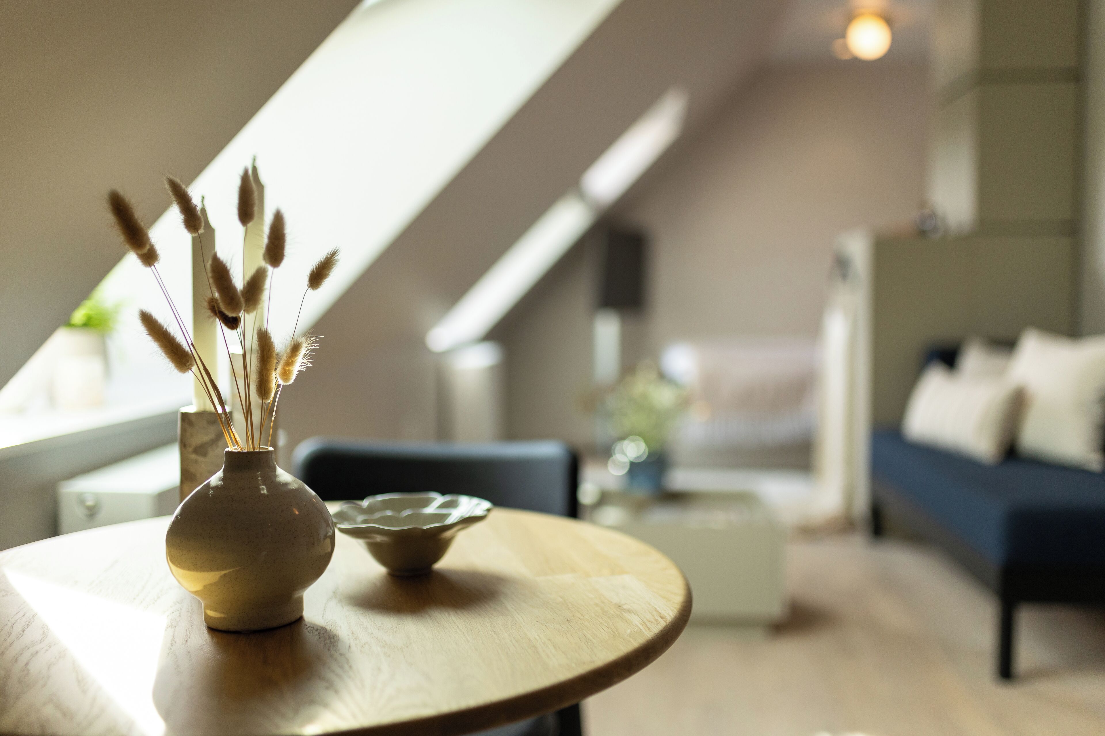 A modern living room with a round wooden table, a vase with dried plants, and a sofa in the background.