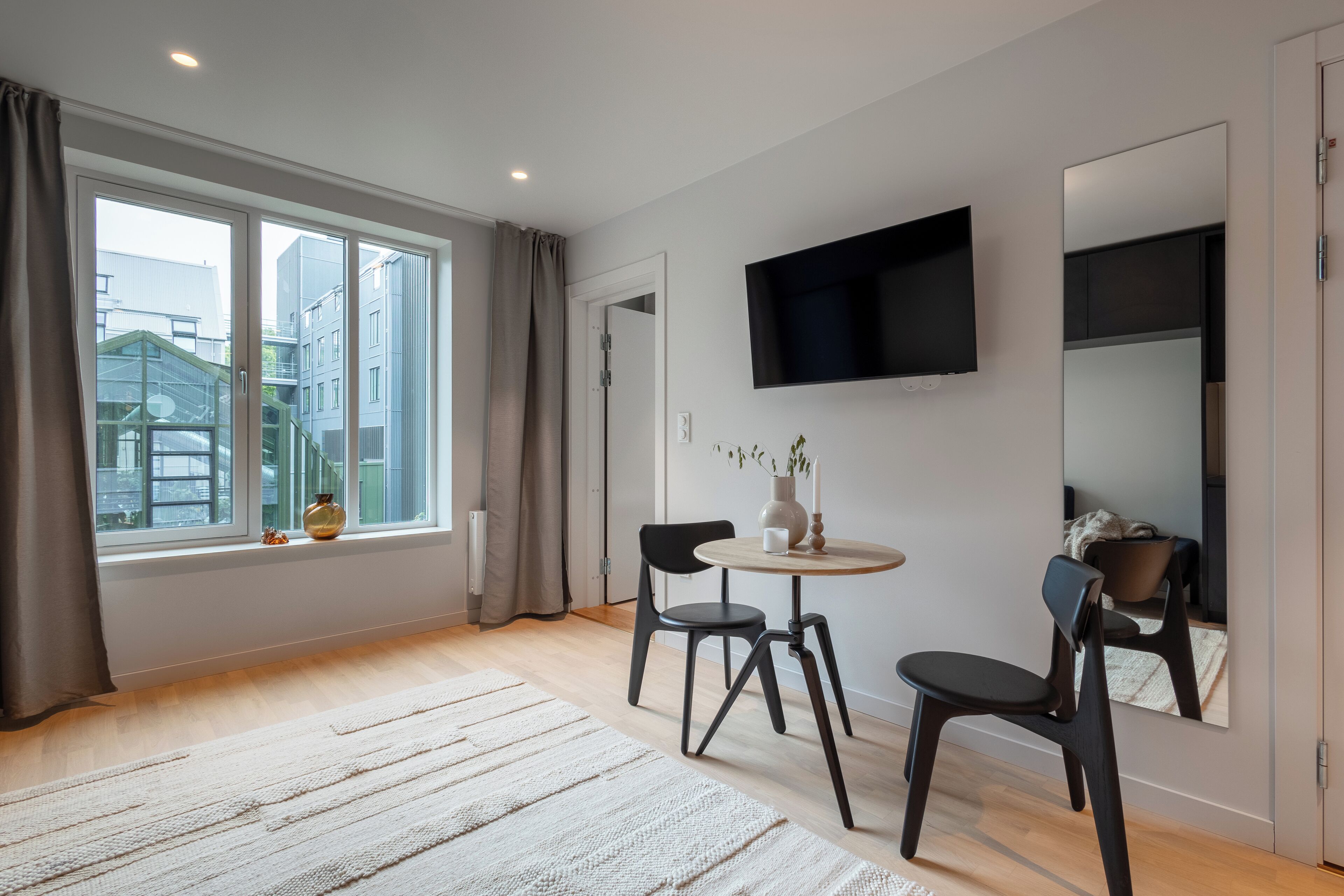 A modern room with a large window, a round table, two chairs, a flat-screen TV, and a mirror.