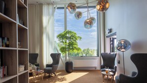 A modern living room with a large window, a potted tree, and a bookshelf. - Unity Malmö - A Studio Hotel (Malmö)