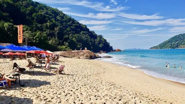 A beach with umbrellas and lounge chairs, a rocky shoreline, and clear blue water. - Raridade - Apt próx praia vista mar ar con (Ubatuba)
