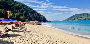 A beach with umbrellas and lounge chairs, a rocky shoreline, and clear blue water. - Raridade - Apt próx praia vista mar ar con (Ubatuba)