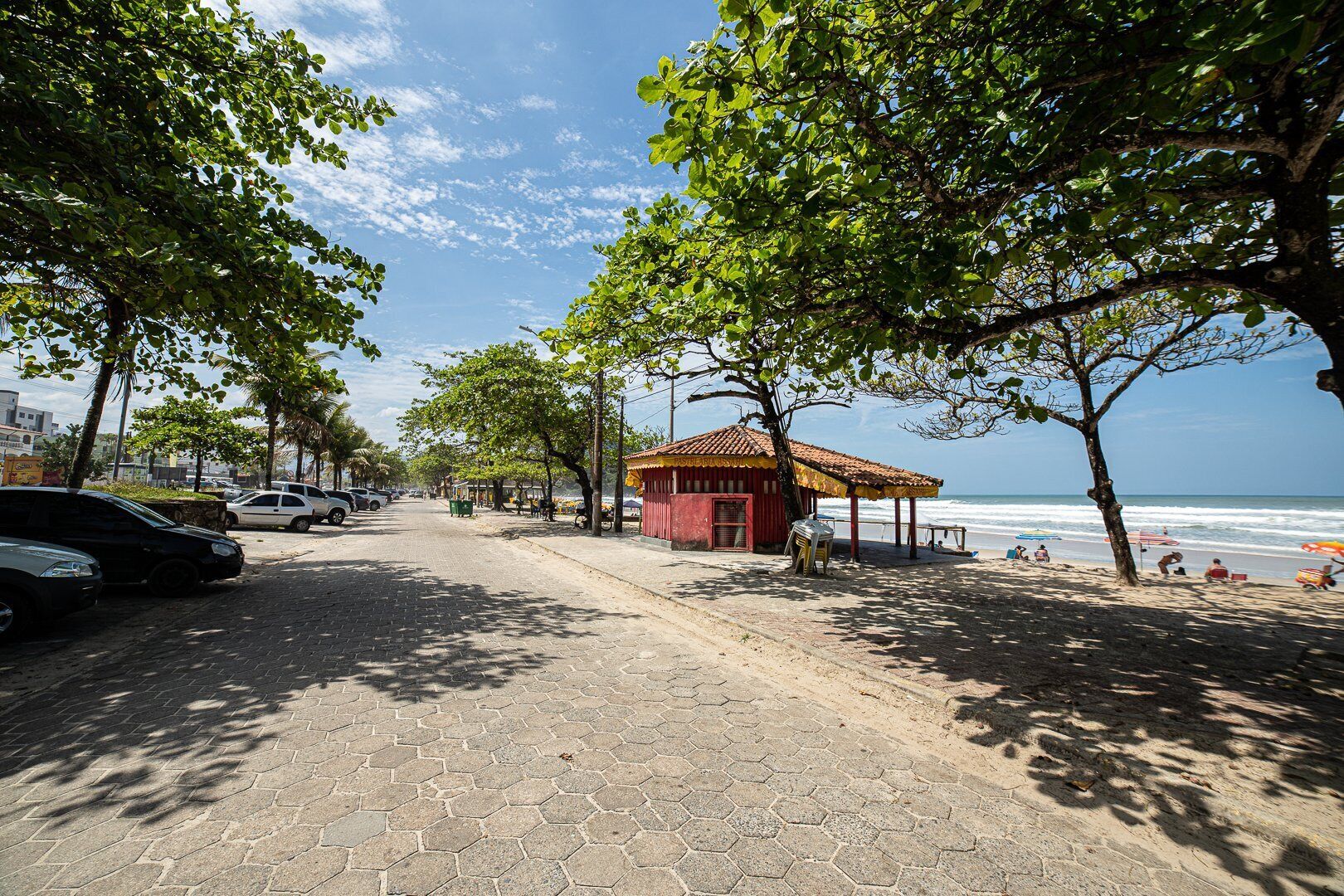 A paved walkway lined with trees, a small red building, and a beach with people in the background.