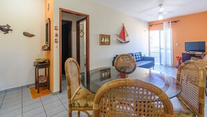 A living room with a glass-top dining table, wicker chairs, a blue sofa, and a television. - Raridade - Apt próx praia vista mar ar con (Ubatuba)