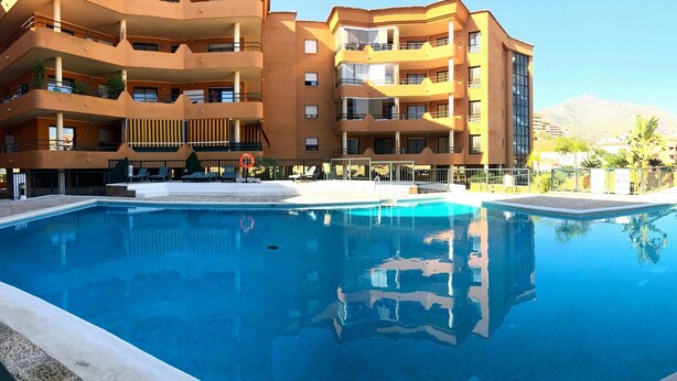 A swimming pool with a clear blue water surface, surrounded by a multi-story building with balconies, and a mountainous backdrop. - Sun Hill Los Pacos 2 Bed Apt (Fuengirola)