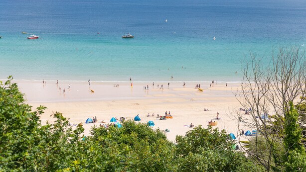 A beach with tents and people, a boat in the water, and a distant island. - Waters Edge (St Ives)
