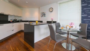 A modern kitchen with white cabinets, a central island, and a dining area with a glass table and grey chairs. - Waters Edge (St Ives)