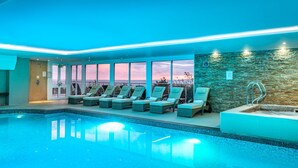 An indoor swimming pool with lounge chairs, a stone wall, and a view of the sunset. - Waters Edge (St Ives)