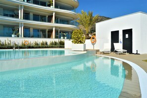 Un hotel moderno con piscina, asientos al aire libre y un edificio blanco con persianas negras.
