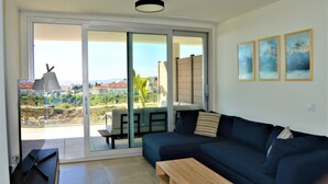 Un salon moderne avec un canapé bleu foncé, une table basse en bois et une porte-fenêtre coulissante donnant sur un balcon avec vue sur la ville.