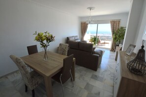 A living room with a sofa, a dining table with chairs, a chandelier, and a view of the sea through a balcony.