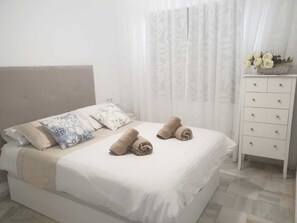 Ein ordentlich bezogenes Bett mit weißer Daunendecke, zwei aufgerollten Handtüchern und Kissen mit floralem Muster.