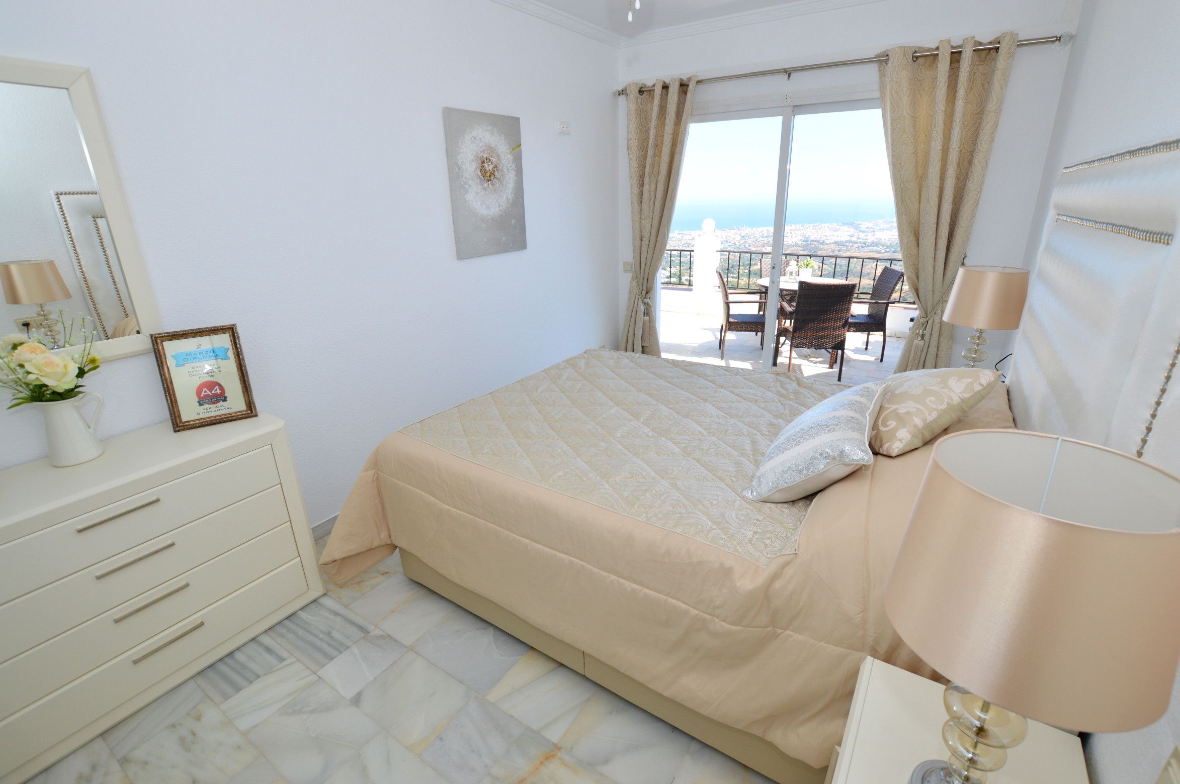 Une chambre avec un lit, une commode, une lampe et une vue sur la ville, visible à travers les rideaux du balcon.