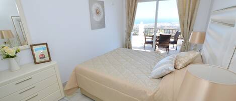 Une chambre avec un lit, une commode, une lampe et une vue sur la ville, visible à travers les rideaux du balcon.